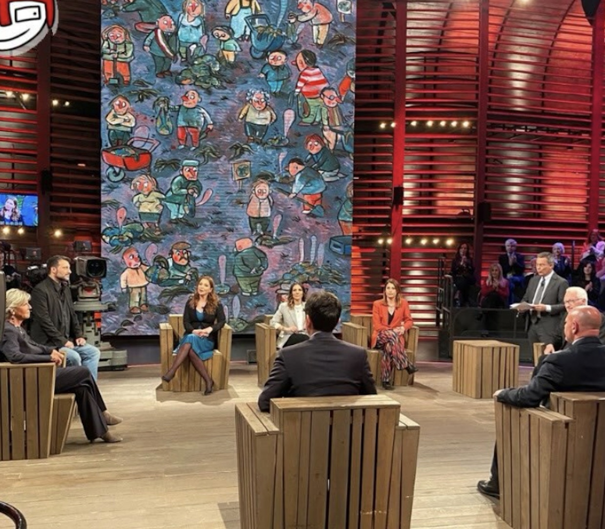 Studio televisivo con grande arazzo dipinto, sedute in legno e illuminazione scenica rossa